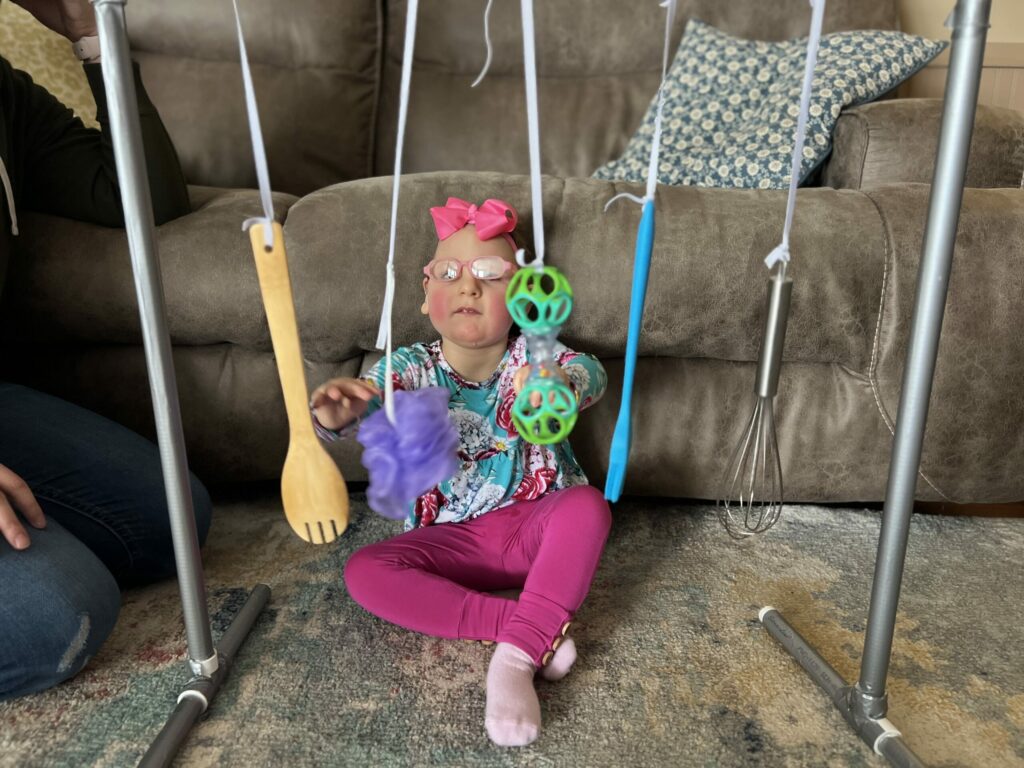 A little girl sitting against the couch playing with kitchen and household items hanging from a play frame. 
