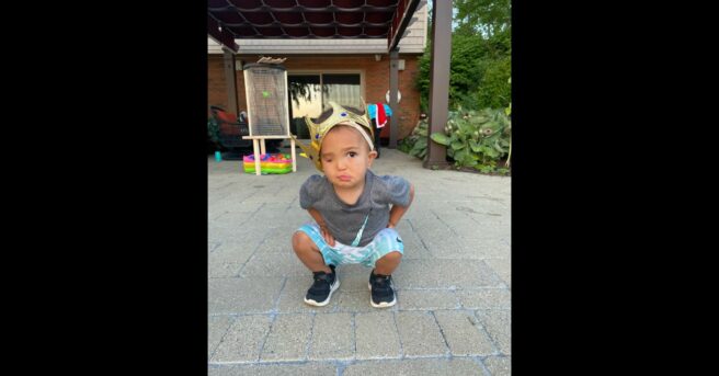 toddler with one eye squats down on a porch while wearing a golden crown