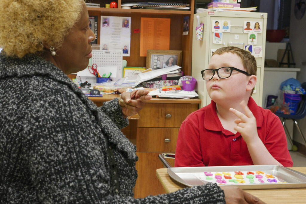 A student using sign to communicate to his teacher sitting next to him.