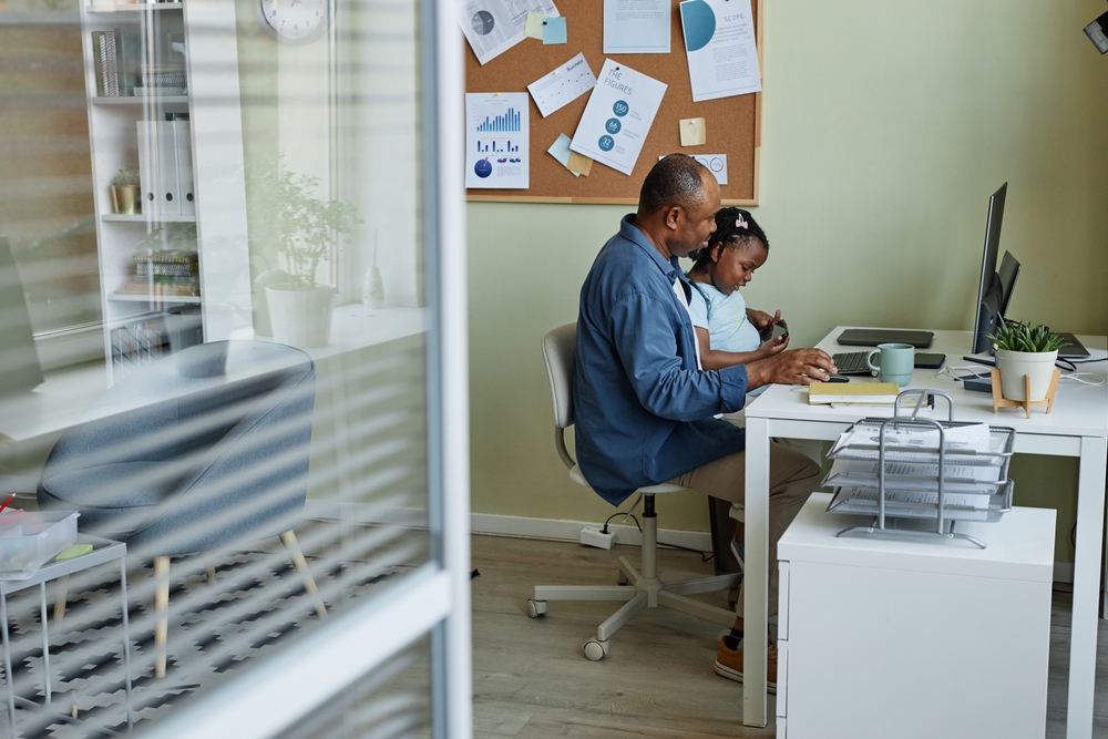 A young girl sitting on her dad's lap working at an office desk.