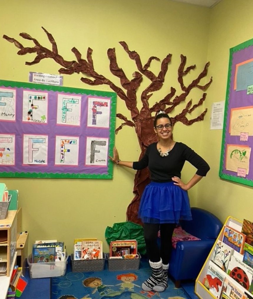 Yokasta Urena is a light skin Latina celebrating Halloween with her Pre-schoolers. She is dressed as a ballerina with her hair up in a bun and wearing a black leotard, Tootoo, and fluffy striped socks. The picture was taken in the classroom library in front of a large paper tree. 