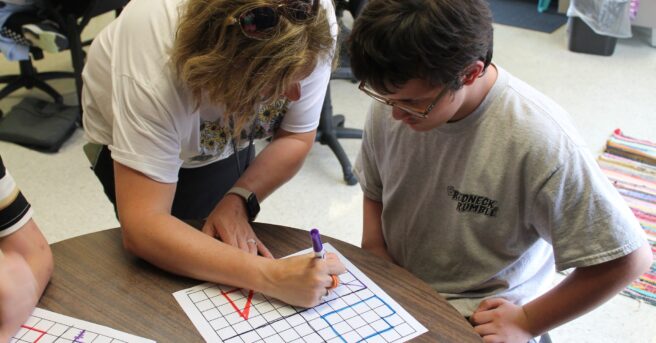 A teacher leaning over to help a student sitting at a table. 