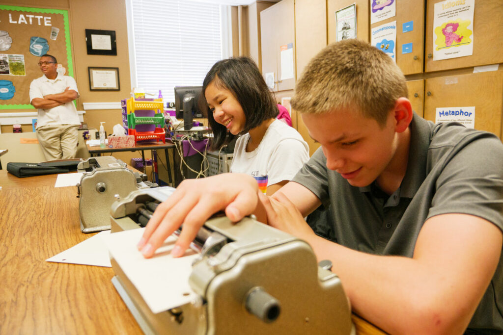 A boy sitting at a table reading braille from his Perkins Braille Writer.