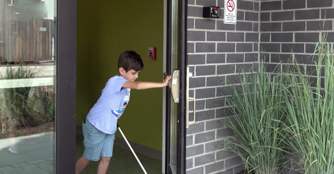 Child opening a door and walking outside while using a white cane
