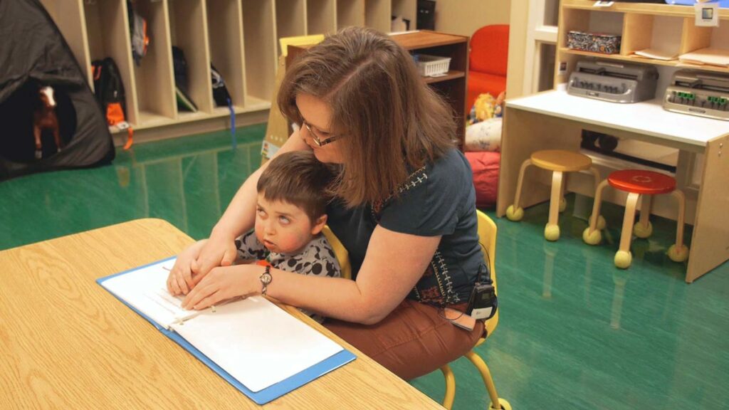 A teacher with her hand on the braille book with the students hand on top of her hand.