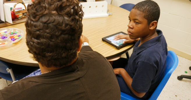 A student working on an iPad with his teacher sitting next to him.
