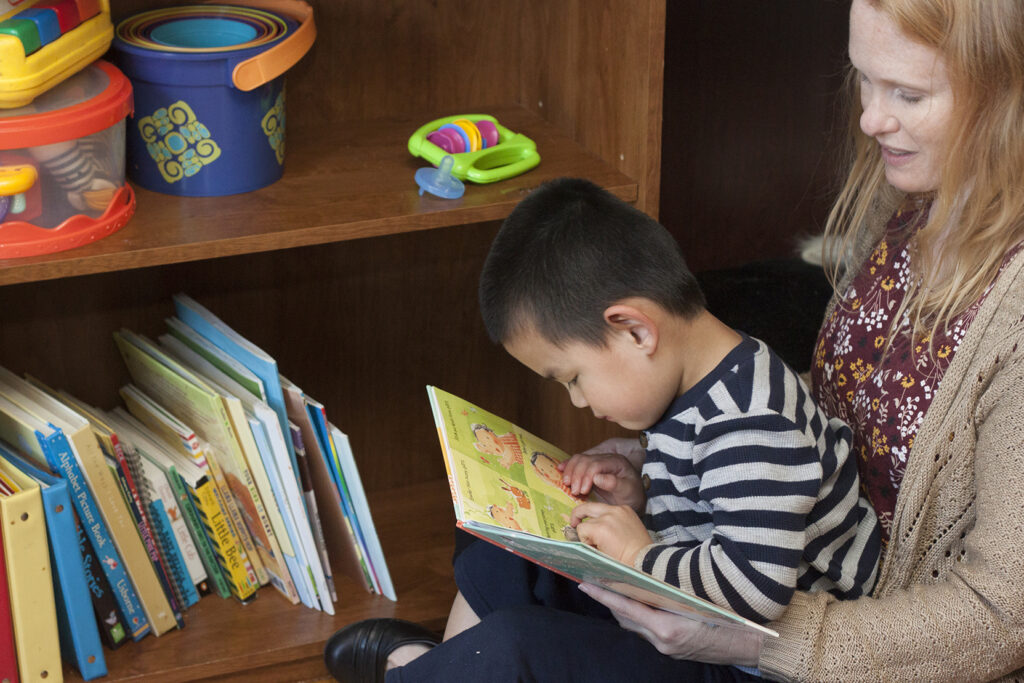 A toddler boy reading a book and feeling braille while sitting on his mom's lap near a book case.