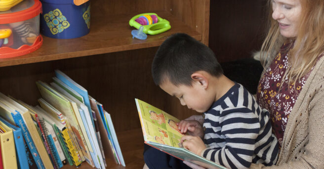 A toddler boy reading a book and feeling braille while sitting on his mom's lap near a book case.