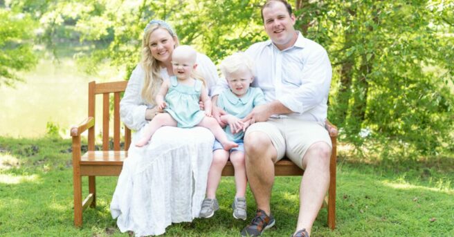 Family with two small children with fair skin and hair sit on bench and smile