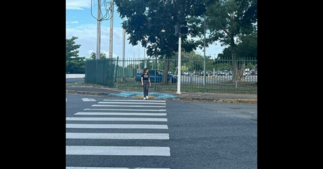 A girl standing in a crosswalk trying to cross. Behind her is an APS that doesn't work. [Una joven intenta cruzar sobre la línea peatonal. Detrás de ella hay un APS que no funciona.]