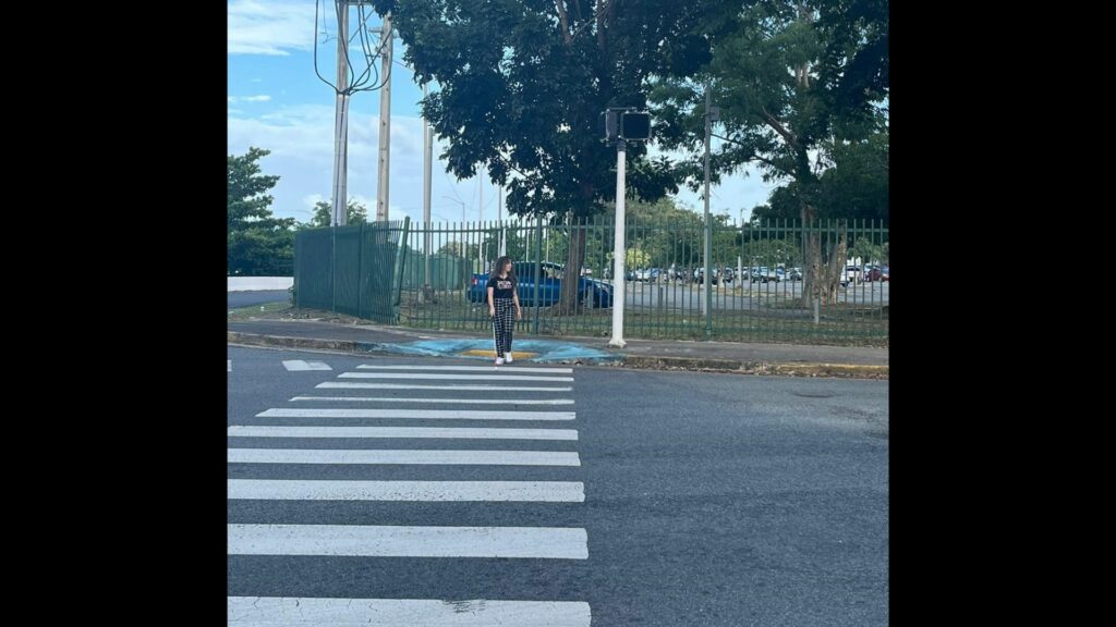 A girl standing in a crosswalk trying to cross. Behind her is an APS that doesn't work. [Una joven intenta cruzar sobre la línea peatonal. Detrás de ella hay un APS que no funciona.]