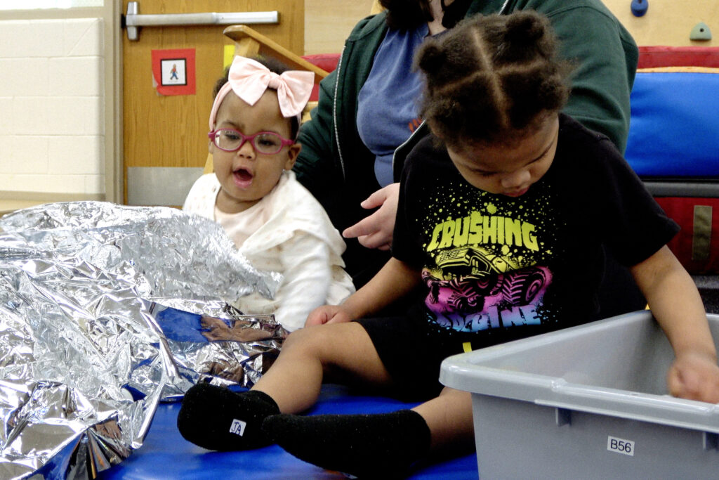 Two children sitting on the floor, one playing with metallic paper and the other playing with silver sound balls.
