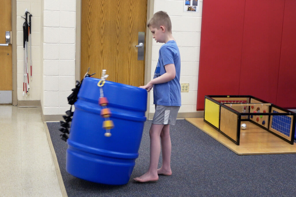 A child standing and engaged with a large blue barrel with bells moving it around.