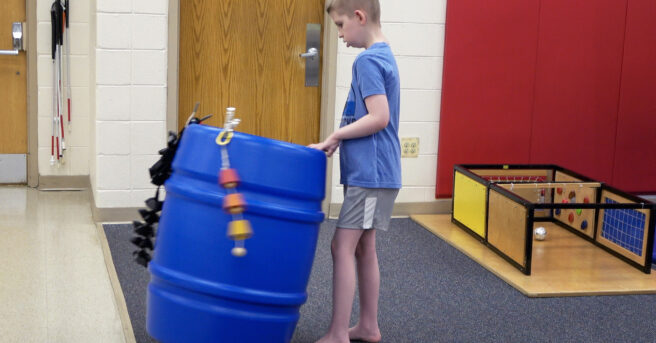 A child standing and engaged with a large blue barrel with bells moving it around.