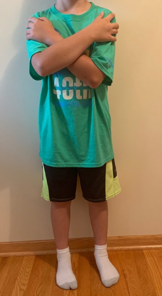 This picture is of a forward-facing boy from the shoulders down placing his arms across his or her chest with each hand touching the opposite shoulder. The boy is clothed wearing a t-shirt, shorts, and socks in this picture.
