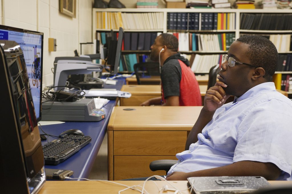 Photo of a young man looking at a computer screen with another individual in the background doing the same.