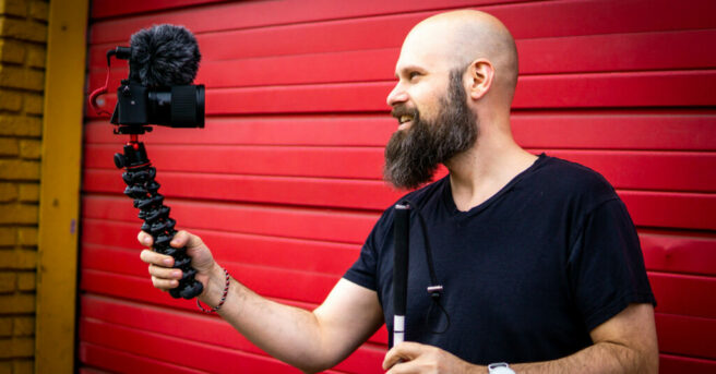 Individual holding a white cane with one hand and a professional camera, directed toward his face, with the other