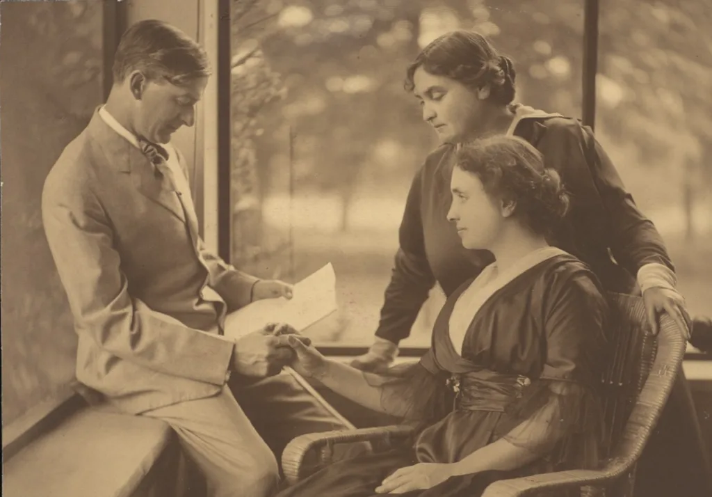 Anne Sullivan stands behind Helen Keller, who is seated and facing slightly right, while John Macy sits on the window ledge to the left, gently spelling into Helen’s hand as she listens attentively by touch.