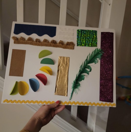 A sensory board with affixed textured papers, ribbons, sandpaper, feathers, shiny fabric, and Velcro.