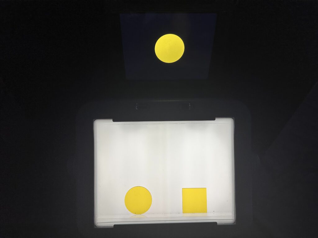 The first photo shows a transparent yellow square and circle sitting on a lightbox with a ledge in a dark room. A yellow circle with a black background displayed on an iPad appears to float behind the lightbox. The iPad is in a black case on top of a black box with a black felt tri-fold board behind it. All of these black materials become invisible in a dark room. 