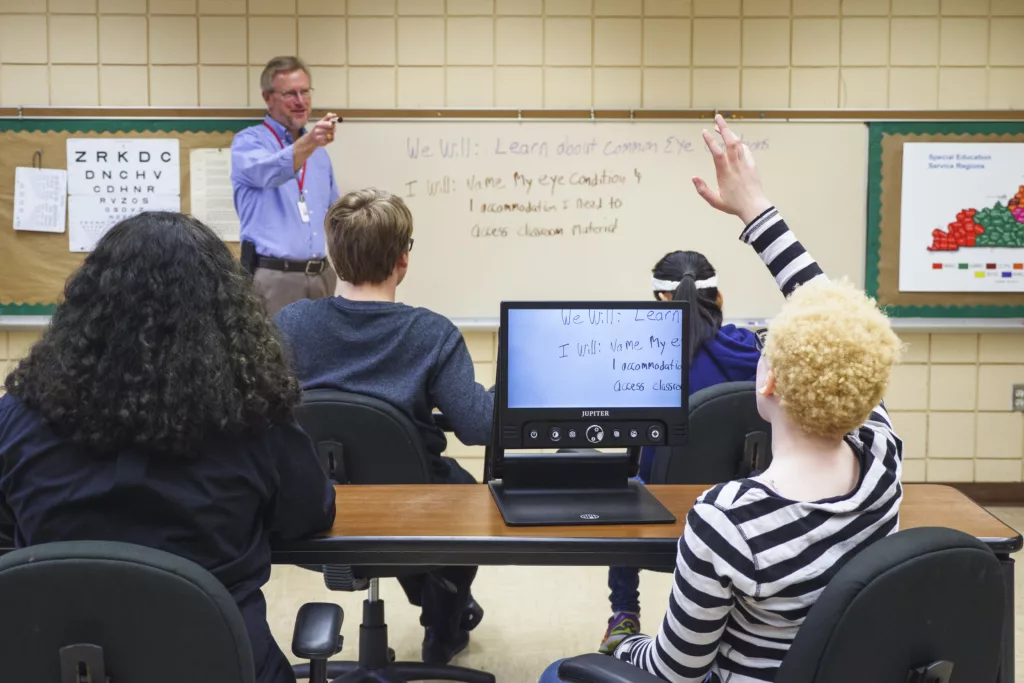 A student raising their hand using a CCTV to see the board, the teacher is pointing at the student.