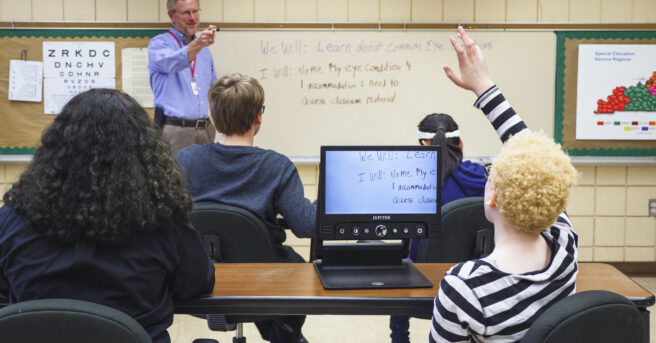 A student raising their hand using a CCTV to see the board, the teacher is pointing at the student.