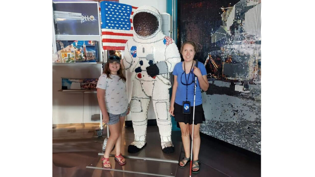 Two teens pose on either side of an astronaut made of Lego