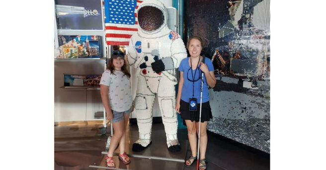Two teens pose on either side of an astronaut made of Lego