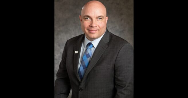 Person in a dark suit with a white shirt and a patterned blue tie smiles for the camera.