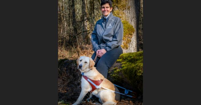 Person stands outside with yellow lab in harness.