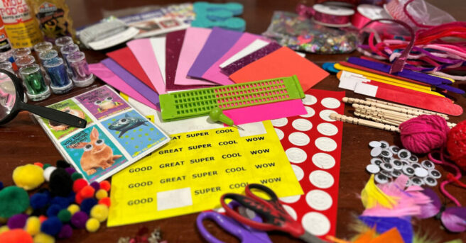 A variety of craft materials displayed on a table. Items include bold print and tactile stickers, a slate and stylus, scissors, store-bought valentine cards, feathers, yarn, google eyes, fuzzy pom-poms, craft sticks, pipe cleaners, ribbon, glitter, and glue.