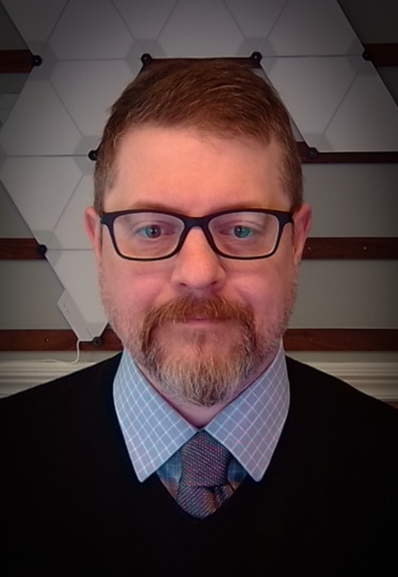 man with close cropped blond hair, brown rimmed glasses and a goatee wearing a button down blue shirt and tie with a black sweater.