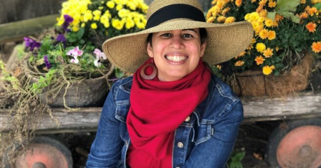 Yokasta Urena is a light skin Latina wearing a large blue hat and blue jean jacket sitting in front of an old wagon with some colorful fall flowers!