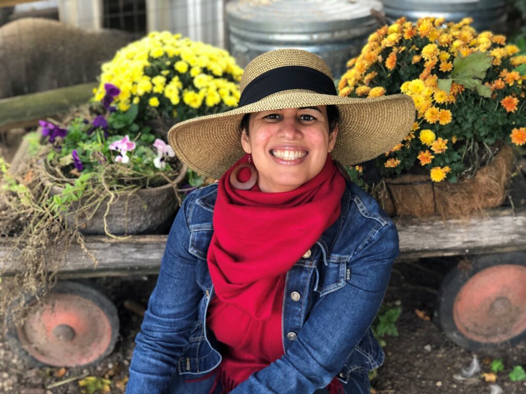 Yokasta Urena is a light skin Latina wearing a large blue hat and blue jean jacket sitting in front of an old wagon with some colorful fall flowers!