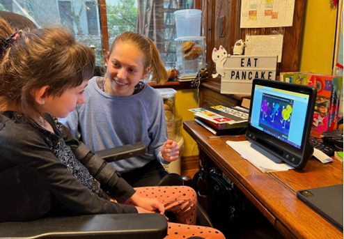 A child sitting in a wheelchair with Adele next to her supporting her learning on a larger tablet.