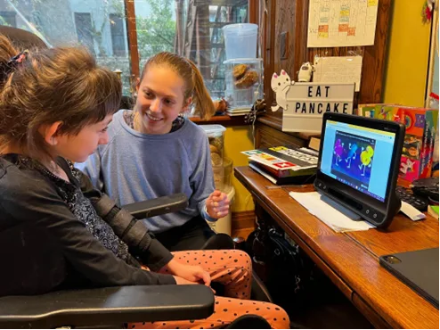 A child sitting in a wheelchair with Adele next to her supporting her learning on a larger tablet.