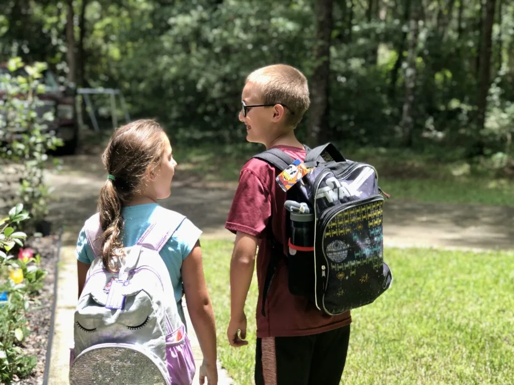 Two children wearing backpacks are walking down a sidewalk, smiling and talking. A taller boy is on the right and a shorter girl is on the left.