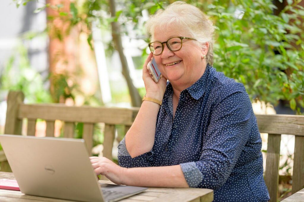 older person sits outside and uses smartphone and laptop for virtual learning