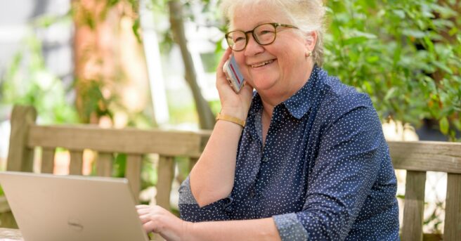 older person sits outside and uses smartphone and laptop for virtual learning