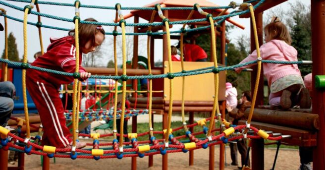 Children playing on a rope bridge and a fort outside at a playground.