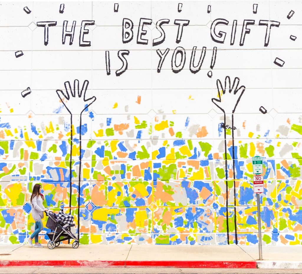 picture of mural on wall with two hands pointing to words "The Best Gift is You!" in the lower foreground is a woman with a stroller walking by. Photo by Dakota Corbin on Unsplash