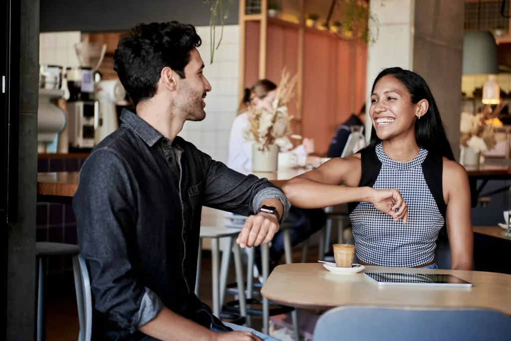 couple sit at a restaurant with espresso while laughing and playfully giving an elbow bump.