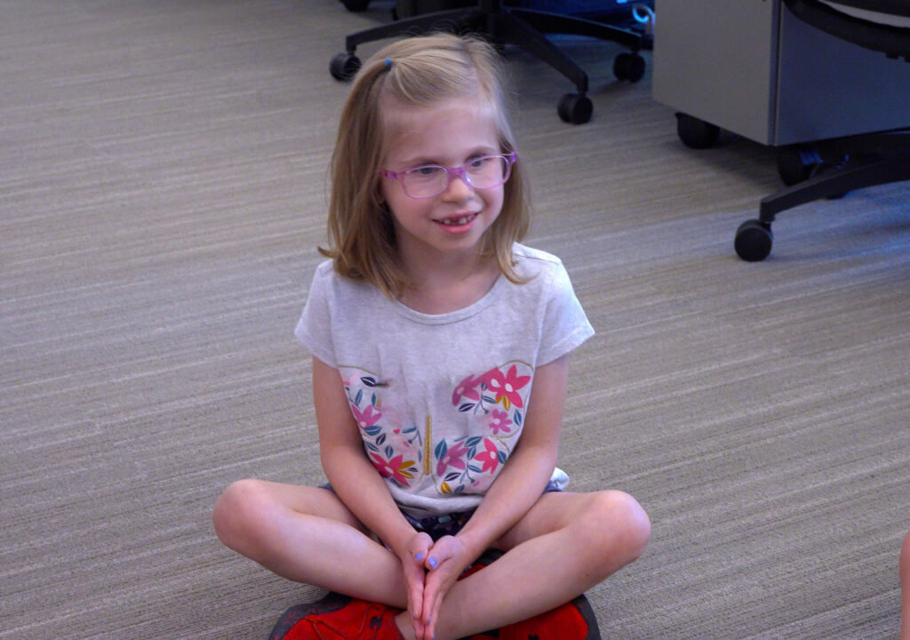 A girl wearing glasses sitting on the floor in a classroom.