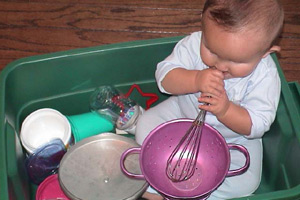baby sitting in a plastic tub holding a whisk,  surrounded by other kitchen items
