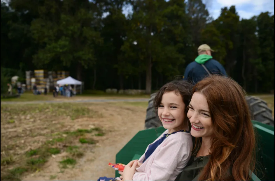 A woman is holding her daughter in the back of a wagon pulled by a man driving a tractor on a country road.