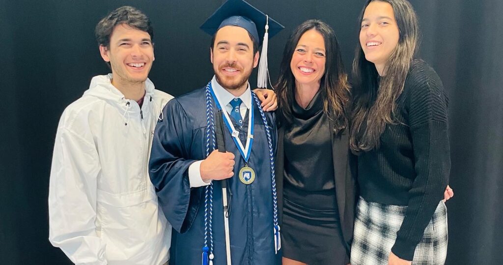 Kristin standing with her three children, Michael, Mitchell, and Karissa. Michael is in a blue Penn State graduation cap and gown, with cords and a medal around his neck, holding his white cane. All have very big smiles.