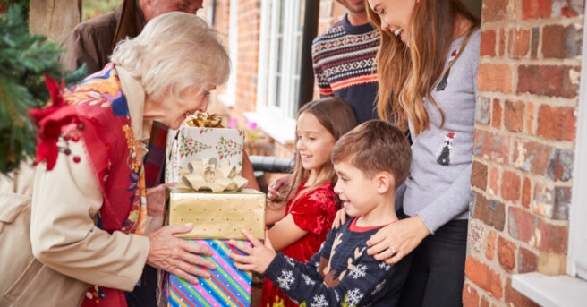 Grandparents are presenting gifts to a mother, father and their two children at the door to their home.