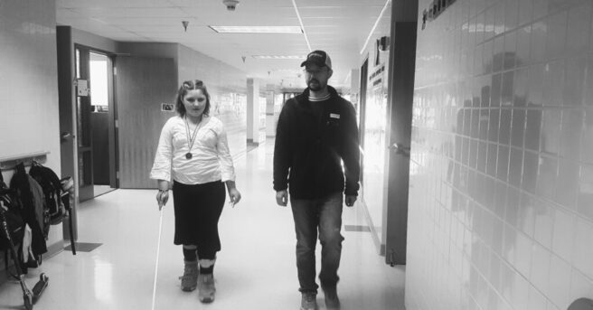 A teen using a white cane and an adult walk down a hallway.