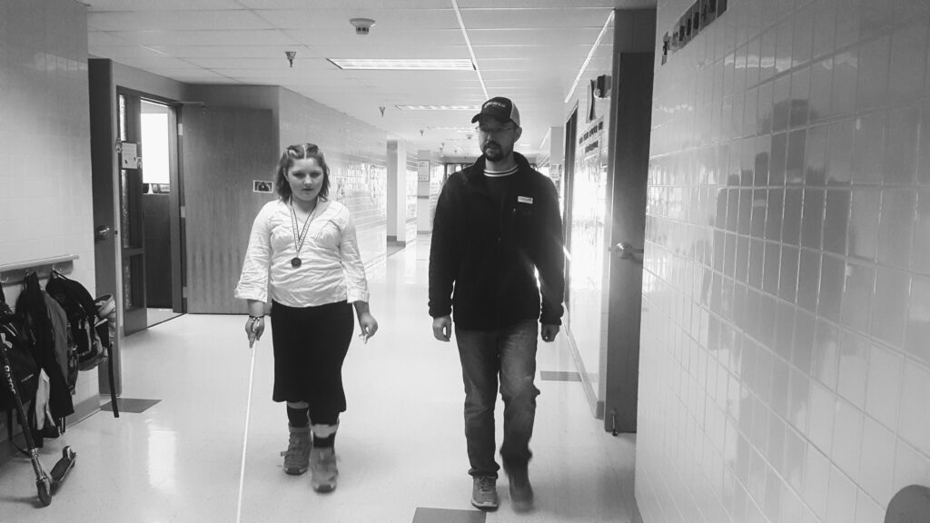 A teen using a white cane and an adult walk down a hallway.