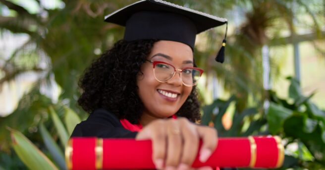 young person with curly hair and eyeglasses wears a graduation cap and gown